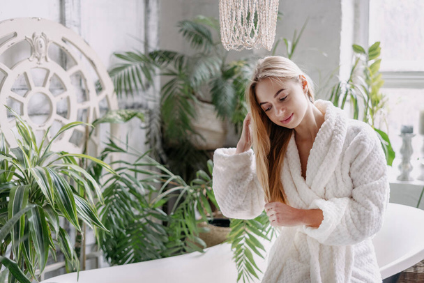 Beauty concept. Young smiling woman in white bathrobe touching hair, standing in light cozy bathroom with green house plants near wall and perform daily haircare routine - Photo, Image