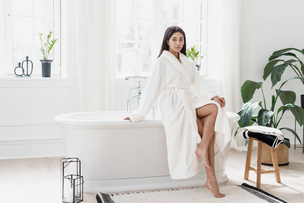 Attractive young asian woman sitting in light spacious bathroom with green plants wearing white bathrobe and showing legs after pampering morning routine. Selfcare concept - Photo, Image