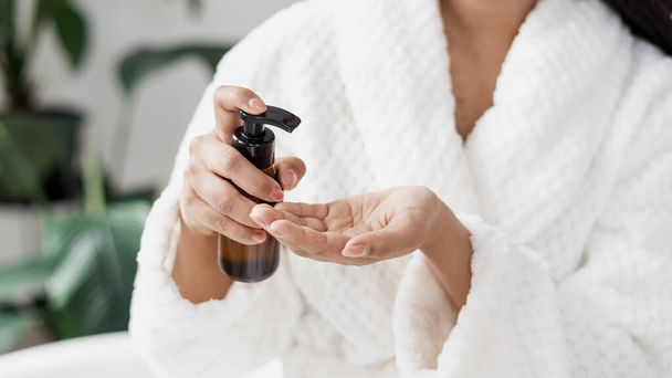 Skincare and natural beauty concept. Selective focus on glass bottle with dispenser in woman hands. Cropped view of young female in bathrobe using organic lotion or body care oil, close up - Foto, afbeelding