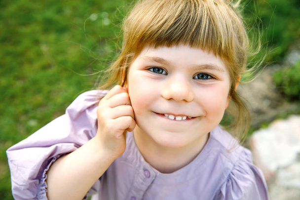 Portrait of happy smiling toddler girl outdoors. Little child with blond hairs looking and smiling at the camera. Happy healthy child enjoy outdoor activity and playing. - Photo, Image