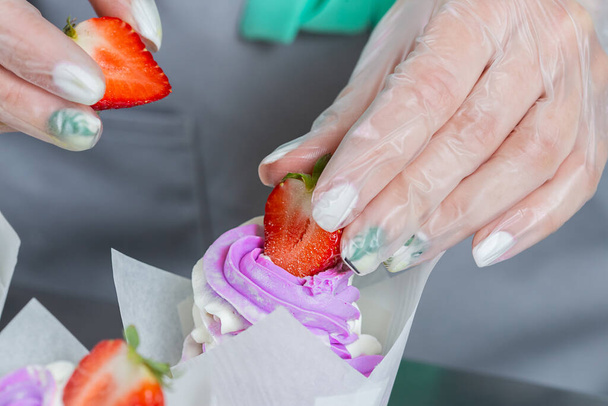 Woman hands chef decorating Cupcakes with pink butter cream and fresh berries in white baking parchment cases on metal table in a professional confectionery studio. - Photo, Image