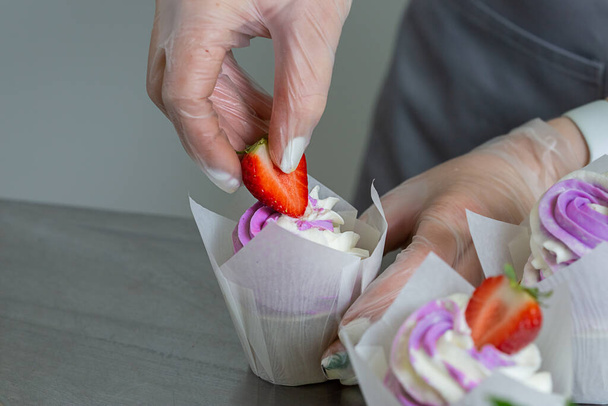 Woman hands chef decorating Cupcakes with pink butter cream and fresh berries in white baking parchment cases on metal table in a professional confectionery studio. - Foto, Bild