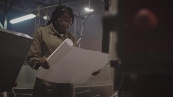 Medium shot of young African female engineer wearing worker overall and safety goggles, holding paper with blueprint, standing in production premises - Footage, Video