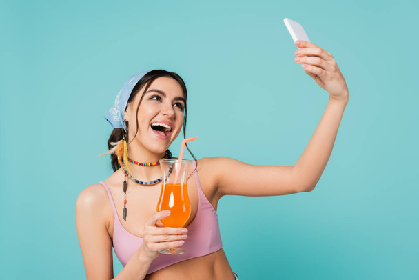 Mulher alegre com coquetel tomando selfie no smartphone isolado no azul  - Foto, Imagem