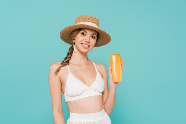 Smiling woman in straw hat holding sunscreen isolated on blue  - Fotoğraf, Görsel