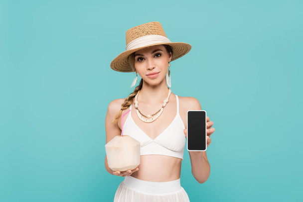 Young woman in sun hat and top holding smartphone and cocktail in coconut isolated on blue  - Foto, imagen
