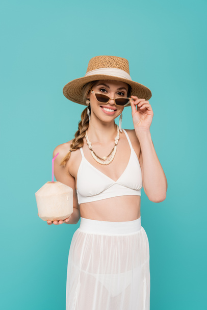 Positive woman in sun hat and swimsuit holding cocktail in coconut isolated on blue  - Fotografie, Obrázek