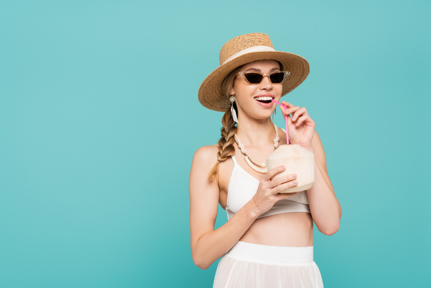 Cheerful woman in sunglasses holding cocktail in coconut isolated on blue  - Photo, Image