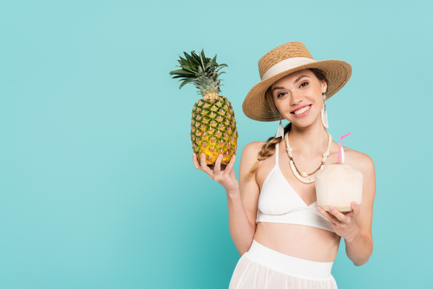 Pretty woman in sun hat holding pineapple and cocktail in coconut isolated on blue  - Fotografie, Obrázek