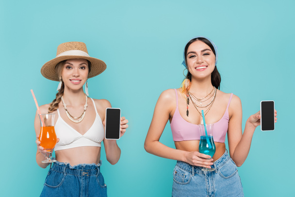 Mujeres sonrientes con tops y collares sosteniendo teléfonos inteligentes y cócteles aislados en azul  - Foto, imagen