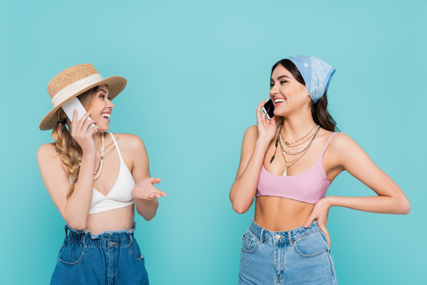 Positive women in tops talking on smartphones isolated on blue  - Foto, Imagen