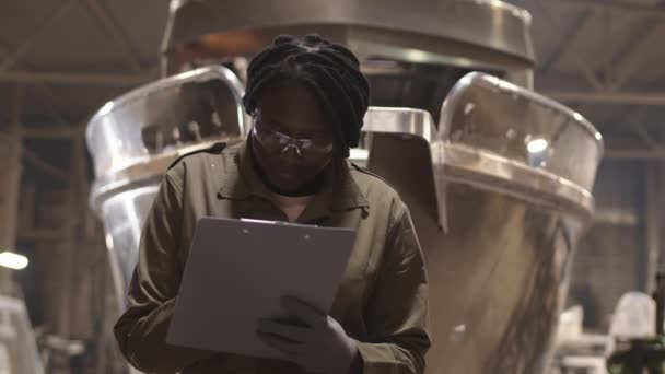 Aufstehen einer jungen afrikanischen Werftmanagerin mit Schutzbrille und Handschuhen, Klemmbrett in der Hand, Blick in die Garage und Notizen machen - Filmmaterial, Video