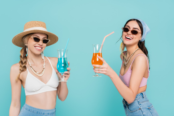 Pozytywne kobiety w topach i okularach przeciwsłonecznych trzymające koktajle odizolowane na niebiesko  - Zdjęcie, obraz
