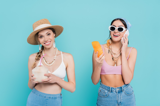 Smiling woman holding cocktail in coconut near friend with sunscreen talking on smartphone isolated on blue  - Фото, изображение
