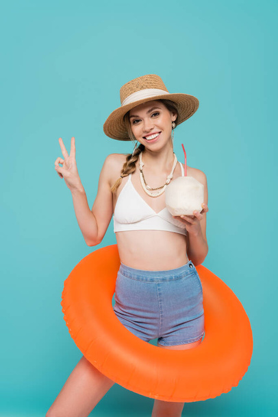 Positive woman in swim ring holding cocktail in coconut and showing peace gesture isolated on blue  - Photo, Image