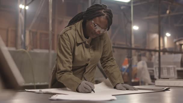Μέτριο πλάνο της γυναίκας Αφρικανή μηχανικός φορώντας φόρμες, γάντια και γυαλιά ασφαλείας, στέκεται δίπλα στο γραφείο στο βιομηχανικό γκαράζ, κάνει αλλαγές στο σχέδιο, χαμογελώντας - Πλάνα, βίντεο