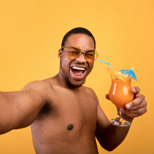 Podekscytowany czarny facet z nagim tułowiem pijący świeże koktajle owocowe i robiący selfie na żółtym tle studio - Zdjęcie, obraz