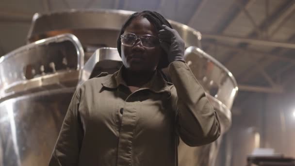 Niski kąt portretu młodej afrykańskiej pracowniczki stoczni stojącej na pierwszym planie metalowej łodzi, zdejmującej okulary ochronne i patrzącej na kamerę - Materiał filmowy, wideo