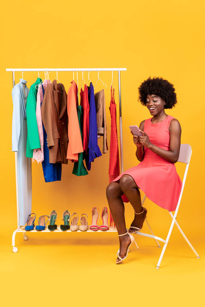 Positiva dama afroamericana usando el teléfono celular comprando ropa, fondo amarillo - Foto, Imagen
