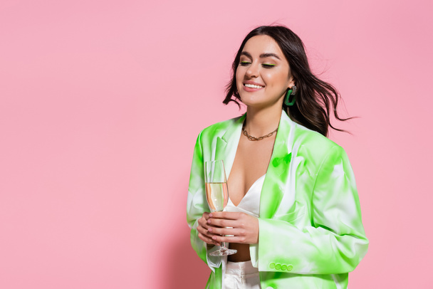 Positive woman in necklace and jacket holding glass of champagne on pink background  - Foto, Imagem