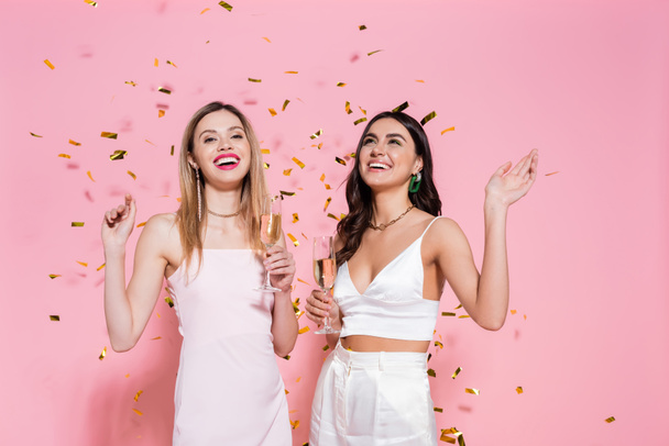 Positive brunette woman with champagne standing near friend and confetti on pink background  - Photo, Image