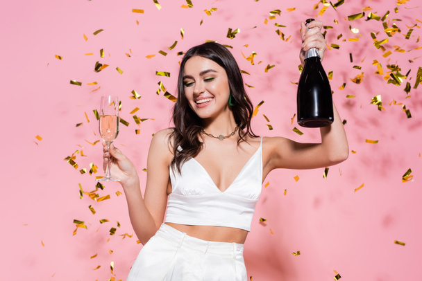 Pretty brunette woman holding bottle and glass of champagne under golden confetti on pink background  - 写真・画像