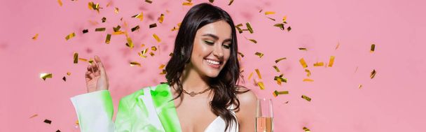 Mujer bonita sonriendo mientras mira la copa de champán cerca de confeti sobre fondo rosa, pancarta  - Foto, Imagen