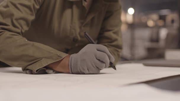 Крупный план неузнаваемого архитектора в комбинезонах и перчатках, наклоняющегося вперед на стол в помещении, вносящего изменения в чертеж - Кадры, видео