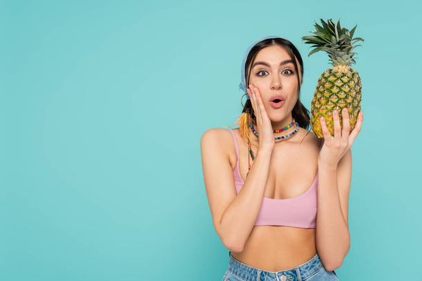 Excited woman in top and necklaces holding pineapple isolated on blue  - Photo, Image