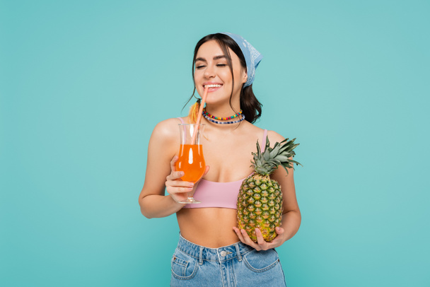 Usmívající se žena pije koktejl a drží ananas izolovaný na modré  - Fotografie, Obrázek
