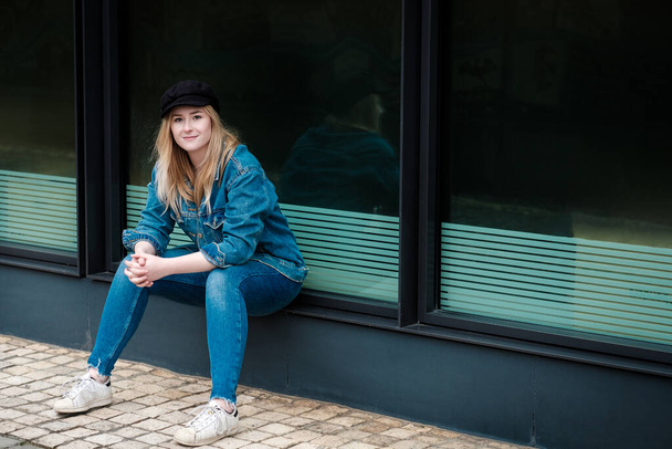 Portrait of young woman sitting on ledge wearing a cap. She is wearing denim trousers and jacket and she is looking to camera. - Фото, зображення