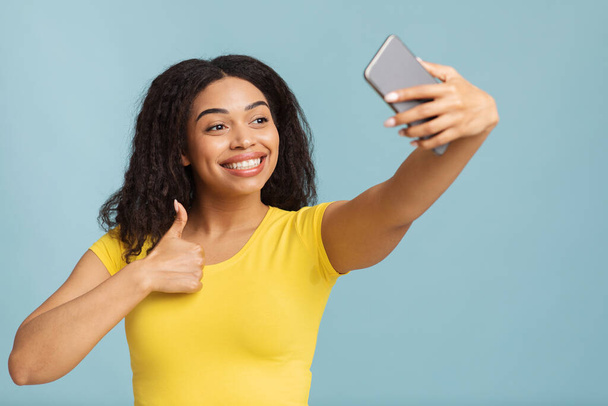 Ich mag es. Glückliche junge schwarze Dame macht Selfie auf Smartphone und zeigt Daumen-hoch-Geste auf blauem Hintergrund - Foto, Bild