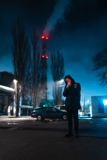 Young man wear coat with hood at the night in neon colored city smokes cigarette - Photo, Image