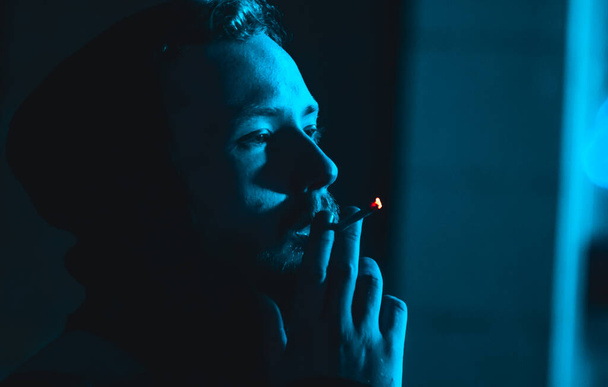 Porträt eines rauchenden gutaussehenden Mannes mit Kapuze bei Dunkelheit mit kaltem blauem Licht  - Foto, Bild