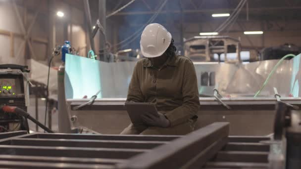 Μέτριο μήκος νεαρής αφρικανής εργάτριας οικοδομών που φοράει προστατευτικά γυαλιά, σκληρό καπέλο και γάντια, κάθεται στην άκρη του μεταλλικού σκάφους, κοιτάζοντας γύρω και χρησιμοποιώντας τον υπολογιστή tablet για να κάνει σημειώσεις - Πλάνα, βίντεο