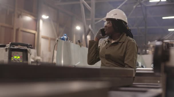 Περίπτωση νεαρής αφρικανίδας οικοδόμου που φοράει προστατευτικά γυαλιά, σκληρό καπέλο και γάντια, κάθεται στην άκρη ενός μεταλλικού σκάφους, μιλώντας στο κινητό τηλέφωνο - Πλάνα, βίντεο