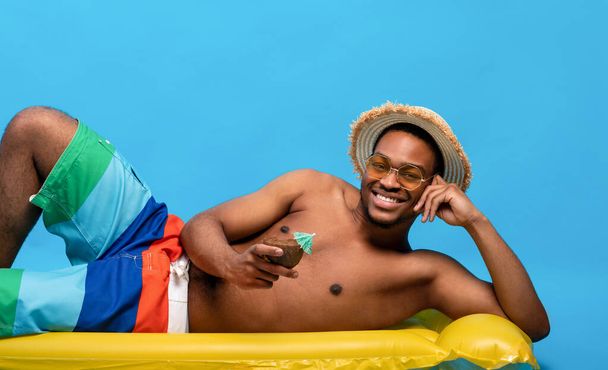 Irrer schwarzer Typ in Badebekleidung liegt auf aufblasbarem Flieder und trinkt sommerlichen Kokoscocktail über blauem Studiohintergrund - Foto, Bild