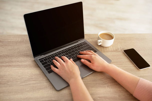 Remote-Arbeitskonzept. Unerkennbare Frau tippt auf Laptop-Tastatur, sitzt mit Tasse Kaffee und Smartphone am Schreibtisch - Foto, Bild