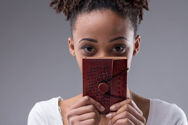Schattige jonge zwarte vrouw staren over een lederen gebonden zuivel ze houdt aan haar gezicht kijken naar de camera met grote ogen in een close-up bijgesneden portret op een grijze studio achtergrond - Foto, afbeelding