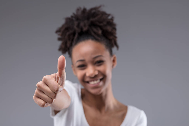 Szczęśliwy uśmiechnięty młody czarny nastolatek dziewczyna daje kciuki w górę gest sukcesu i aprobaty na szarym tle studio z naciskiem na rękę - Zdjęcie, obraz