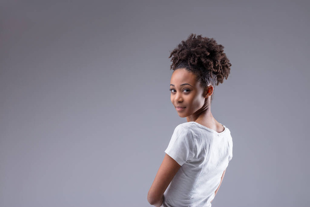 Genç siyahi kadın, sessiz bir gülümseme ve telif hakkı olan gri stüdyo arka planında düşünceli bir ifadeyle omzunun üzerinden kameraya bakıyor. - Fotoğraf, Görsel