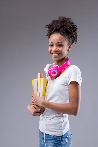 Šťastné živé dospívající dívka drží barevné učebnice v rukou, jak se šklebí na kameru se sluchátky kolem krku na šedém pozadí studia - Fotografie, Obrázek