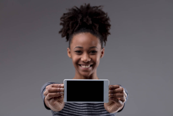 Gelukkig opgewonden jonge zwarte vrouw houden van haar mobiele telefoon naar de kijker met een leeg scherm en focus op de mobiele telefoon op een grijze studio achtergrond - Foto, afbeelding