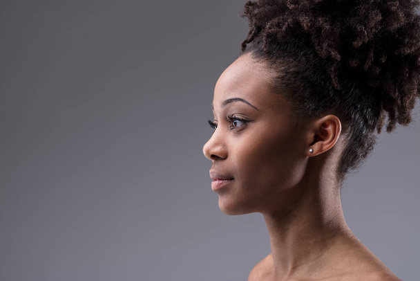 Profilo ritratto di una giovane donna nera seria e bella con i suoi capelli ricci elegantemente legati in cima alla testa guardando verso il copyspace su uno sfondo grigio da studio - Foto, immagini
