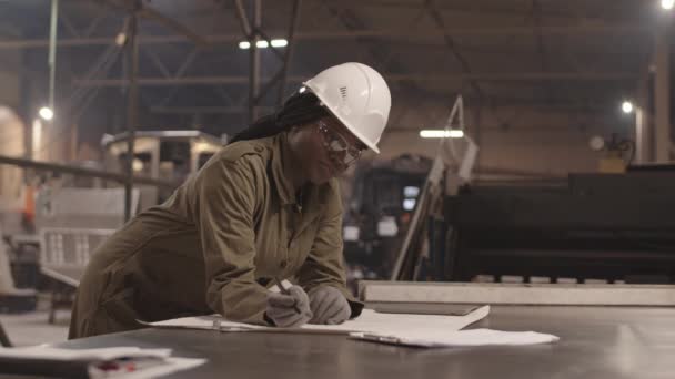 Medium shot of female African engineer wearing overalls, gloves, hard hat and safety goggles, leaning on desk in manufacture, making changes to blueprint - Footage, Video