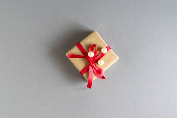 On a gray background, a gift in craft packaging with a red ribbon and white beads. Layout. Gift for Christmas, Valentine's Day, Women's Day, Mother's Day. Horizontal photo. Place for your text. - Photo, Image