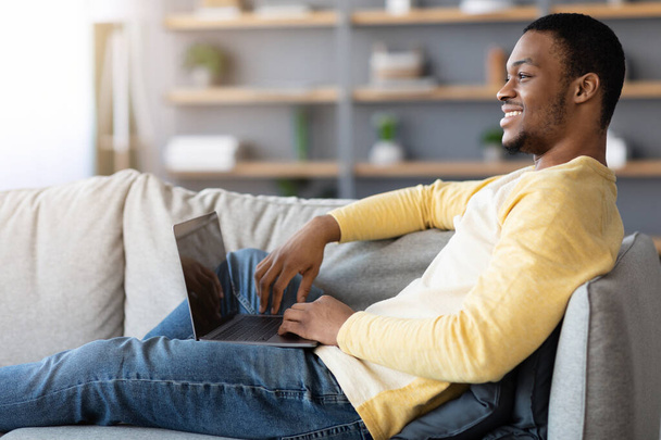 Relaxált fekete srác laptoppal, üres képernyővel, otthoni belső - Fotó, kép