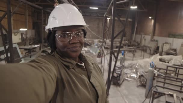 Handgestütztes Brustbild einer jungen afrikanischen Arbeiterin mit weißem Hut und durchsichtiger Schutzbrille, die sie am Arbeitsplatz filmt, winkt und vor der Kamera spricht - Filmmaterial, Video