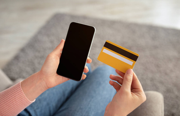 Concetto di shopping online. Donna holdng carta di credito e smartphone con schermo bianco, seduta sul divano di casa - Foto, immagini