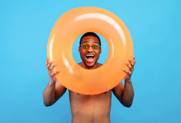 Portrait of excited black guy in sunglasses looking at camera through inflatable ring on blue studio background - Фото, зображення